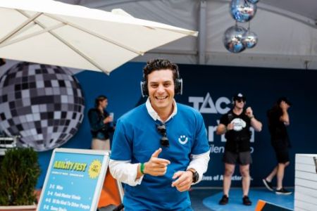 Man with headphones on in blue shirt smiling to camera. He's standing in front of an Ability Fest sign and TAC sign in the background with other people behind listening to music.